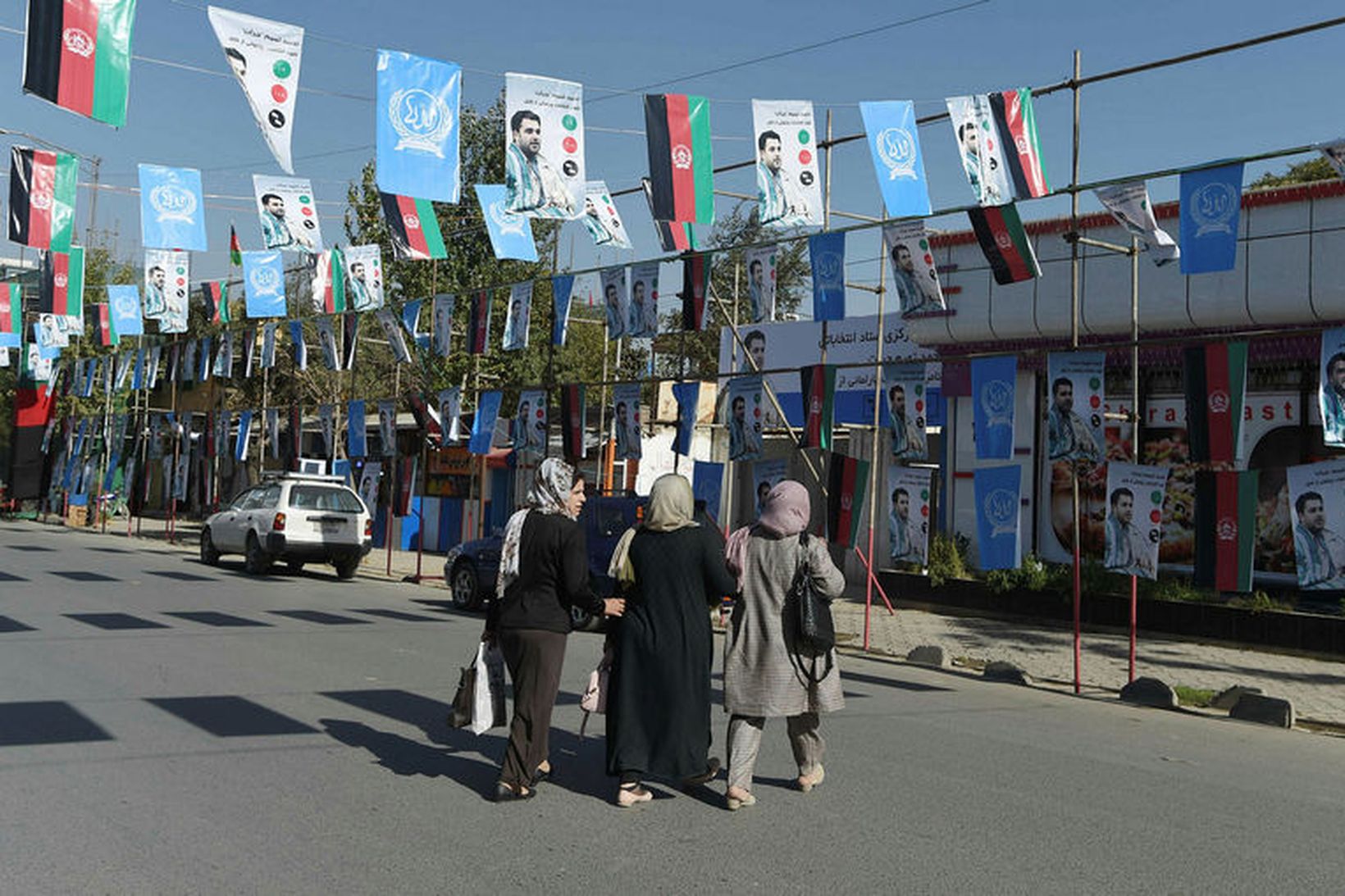 Afganskar konur ganga yfir götu með fjölda plakata af frambjóðandanum …